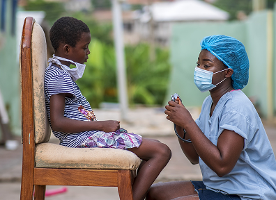 A health worker in a face mask examining a young child seated outdoors
